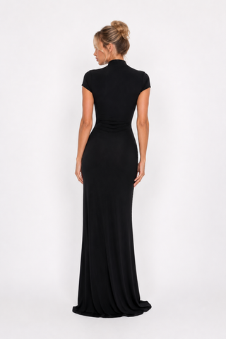 A woman viewed from the back wearing a floor-length black dress with short cap sleeves and a high mock neck, standing against a plain light gray studio background.