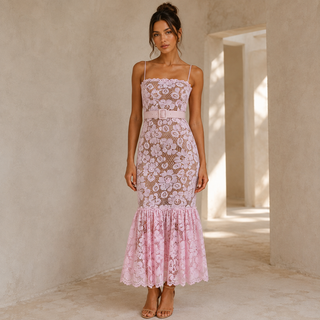 A woman stands in a sunlit architectural setting wearing a fitted light pink floral lace midi dress with thin straps, a matching waist tie, and a flared ruffled hem.