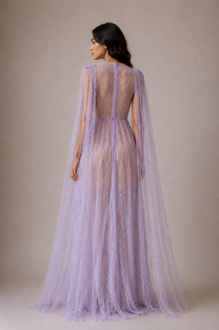 A woman shown from the back wearing a lavender tulle maxi dress with sheer long cape sleeves and a lace-up open back, photographed against a plain warm beige studio background.