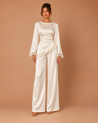 A woman models a cream satin jumpsuit with long bell sleeves, lace trim on the cuffs and waist, and wide legs, set against a solid tan studio backdrop.