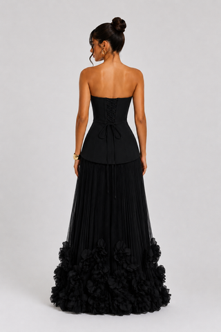 A woman modeling a strapless black gown from the back, featuring a structured corset-style bodice with a bow, a pleated skirt, and a ruffled floral hem, against a light gray studio background.