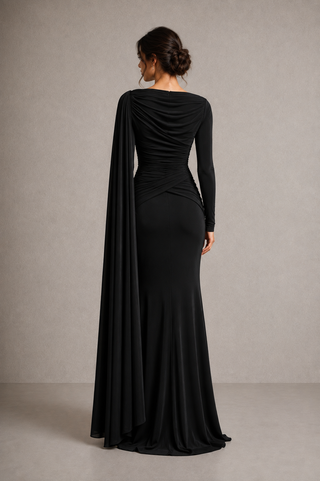 A woman modeling a floor-length black gown from the back, featuring a draped neckline with a long flowing capelet, long sleeves, and a fitted silhouette against a neutral studio backdrop.