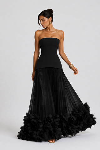 A woman modeling a strapless black gown with a structured corset-style bodice and a voluminous pleated skirt featuring a ruffled tulle hem, photographed against a light gray studio background.