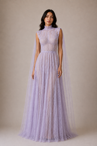 A woman modeling a lavender lace high-neck gown with a sheer tulle cape overlay, set against a solid beige studio background.