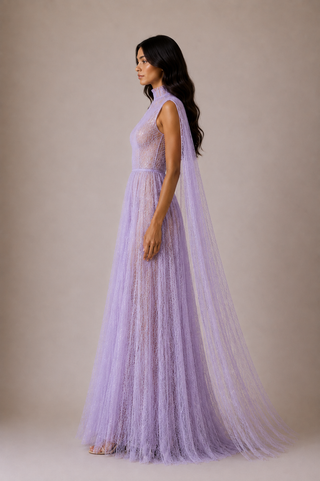 A woman modeling a lavender sleeveless maxi dress with a sheer lace bodice, a full tulle skirt, and a matching long tulle train against a plain beige studio background.
