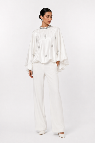 A woman modeling a white two-piece outfit consisting of a long-sleeve blouse with silver embellishments and matching wide-leg trousers, photographed against a seamless white studio background.