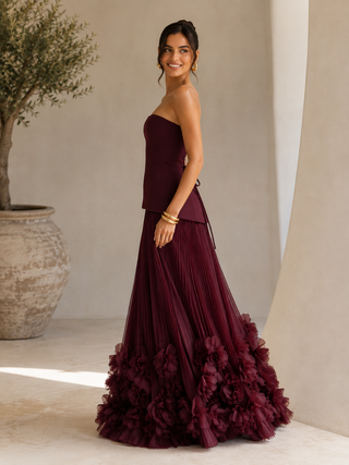 A woman modeling a strapless floor-length gown in deep burgundy. The dress features a structured bodice, a pleated skirt, and a dramatic ruffled floral hem.