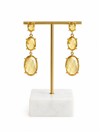 A pair of gold drop earrings with three oval yellow gemstones each, displayed on a gold T-stand with a white marble base against a white background.