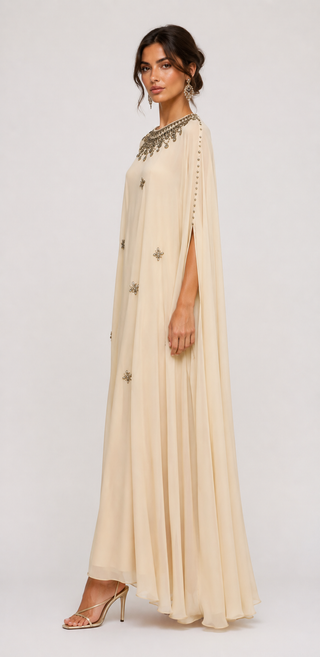 A woman modeling a floor-length cream or beige caftan-style dress with metallic star embellishments and a heavily decorated neckline, photographed against a plain light gray studio background.