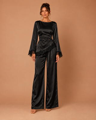 A woman modeling a black satin jumpsuit with wide legs, long flared sleeves, and lace trim at the cuffs and waist, standing against a solid tan studio backdrop.