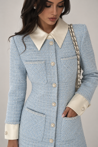 A woman wearing a light blue tweed coat with a contrasting cream satin collar, cream buttoned cuffs, and cream pocket trim, photographed against a neutral studio background.