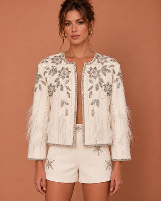 A model wears a cream-colored embellished jacket with silver beadwork, feathered cuffs, and matching cream shorts against a solid terracotta background.