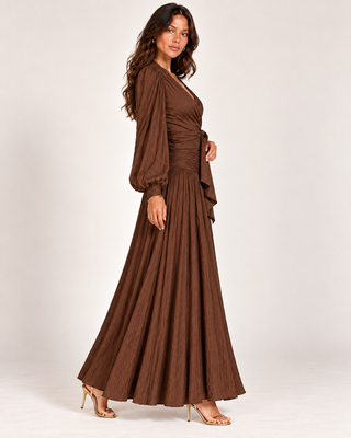 A woman modeling a brown maxi dress with long balloon sleeves, a V-neckline, and a pleated skirt, standing against a light gray studio background.