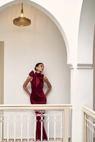 A woman stands on an indoor balcony wearing a sleeveless deep burgundy dress with a large bow at the neckline. The setting features white walls, an arched alcove, a hanging brass pendant light, and white metal railings.