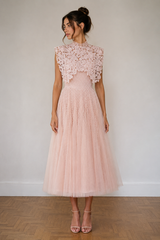 A woman modeling a light pink sleeveless midi dress with a textured lace bodice, high neck, and a voluminous tulle skirt, standing against a neutral off-white wall and wooden parquet floor.