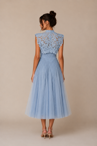A woman viewed from the back wearing a light blue sleeveless dress with a lace high neckline and a full tulle skirt, standing against a plain beige studio background.
