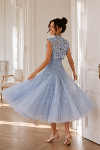 A woman wearing a light blue sleeveless tulle dress with a lace high-neck back, posing in a bright, sunlit room with white walls and wooden floors.