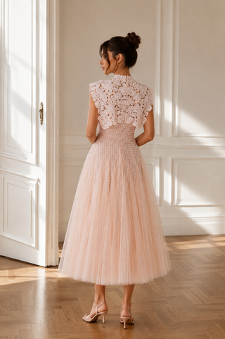 A woman stands in a sunlit room wearing a sleeveless blush pink midi dress with a floral lace back and a full tulle skirt, paired with nude high-heel sandals.