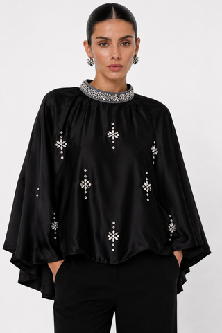 A woman modeling a black satin-like blouse with silver embellished snowflake motifs and a beaded silver collar, paired with black trousers against a light gray studio background.