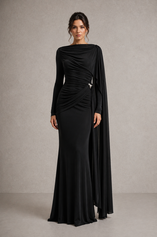 A woman models a floor-length black evening gown with long sleeves, a draped asymmetrical neckline, and a flowing cape-like side drape, standing against a textured light gray studio backdrop.