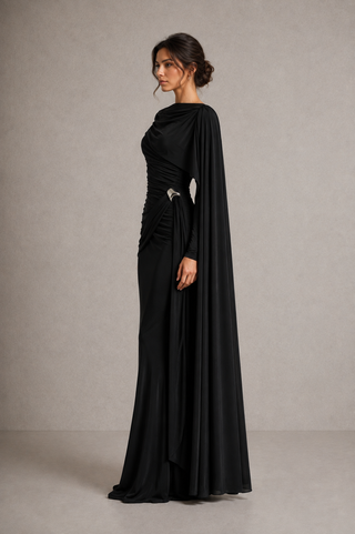 A woman modeling a floor-length black gown with draped fabric, a draped neckline, and a fitted ruched bodice, standing against a plain warm gray studio background.