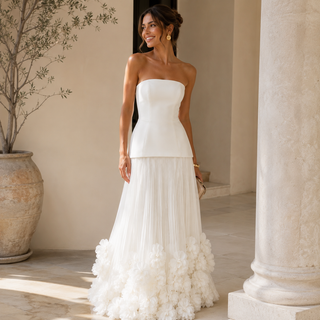 A woman wearing a strapless white wedding dress with a structured bodice and a voluminous ruffled skirt, standing in a sunlit architectural setting with stone columns and a large potted plant.