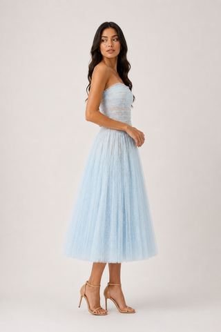 A woman modeling a strapless light blue tulle midi dress with a ruched bodice, paired with nude high-heeled sandals, against a plain light beige studio background.