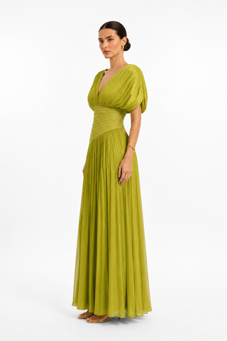 A woman modeling a floor-length olive green gown with a draped V-neck bodice, short puff sleeves, a gathered waist, and a flowing pleated skirt, photographed against a clean white studio background.