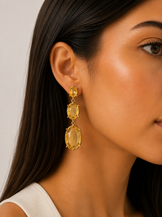 A close-up of a woman wearing a gold drop earring with three stacked oval yellow gemstones, shown against a light neutral background.