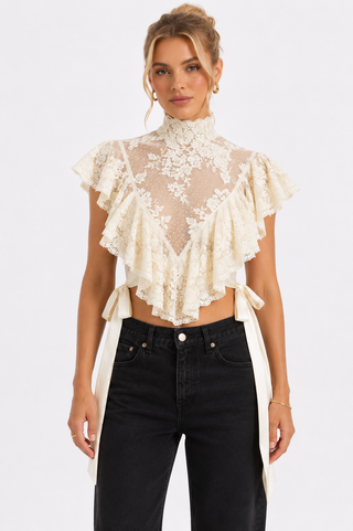 A woman modeling a cream lace mock-neck ruffled crop top with side tie details, paired with black jeans, against a plain light gray studio background.
