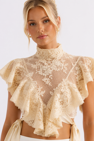 A woman wearing a cream lace ruffled crop top with a high mock neck and side tie details, photographed against a clean white studio background.
