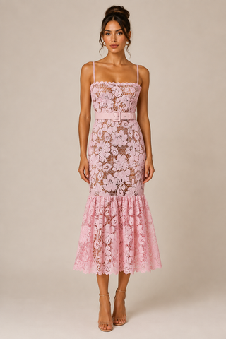 A woman modeling a light pink floral lace midi dress with spaghetti straps, a defined waist, and a flared ruffled hem, set against a solid beige studio background.