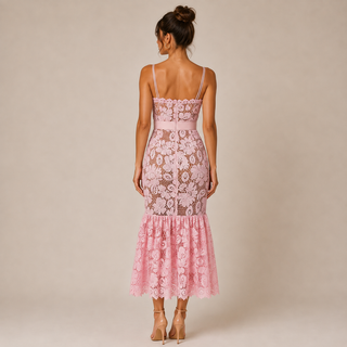 A woman stands with her back to the camera wearing a light pink floral lace midi dress with spaghetti straps and a flared ruffled hem, paired with nude high-heeled sandals, against a solid beige studio background.