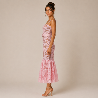 A woman models a light pink floral lace midi dress with spaghetti straps, a matching waist belt, and a flared tulle hem, set against a solid beige studio background.