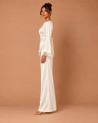 A woman modeling a floor-length white satin robe-style dress with long sleeves, lace trim at the cuffs, and a tie waist, posed against a solid warm tan studio backdrop.