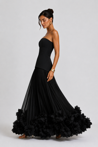 A woman modeling a strapless black formal gown with a structured bodice and a voluminous ruffled tulle skirt, posed against a light gray studio background.