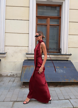 A woman wearing a sleeveless, high-neck, floor-length burgundy dress and black strappy heels walks along a city sidewalk in front of a beige building with a window.