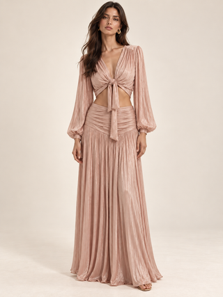 A woman models a dusty pink two-piece set featuring a long-sleeve crop top with a front tie and a matching pleated maxi skirt, set against a plain beige studio background.