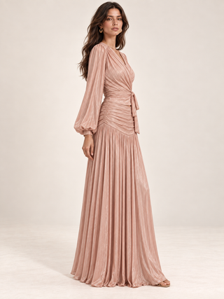 A woman modeling a floor-length pleated gown in a dusty rose pink color, standing against a plain light beige studio background.