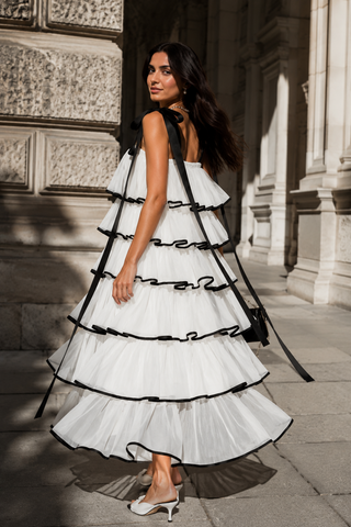 A woman wearing a white tiered tulle dress with black ribbon trim and black shoulder straps, standing outdoors in front of a stone building.