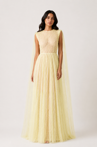 A woman modeling a pale yellow lace and tulle maxi dress with a high neckline and a flowing sheer cape-like skirt, photographed against a plain light gray studio background.