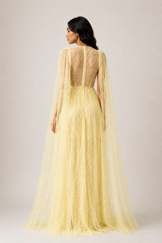 A woman shown from the back wearing a pale yellow lace and tulle evening gown with long sheer cape sleeves and a sheer open back, photographed against a plain light beige studio background.