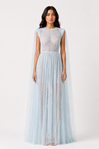 A model wearing a light blue lace and tulle maxi dress with a sheer cape-like overlay, photographed against a plain white studio background.