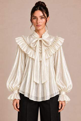 A woman modeling a cream-colored, semi-sheer long-sleeve blouse with dramatic ruffles, a large bow at the collar, and pearl-like decorative accents, paired with black trousers against a solid beige background.
