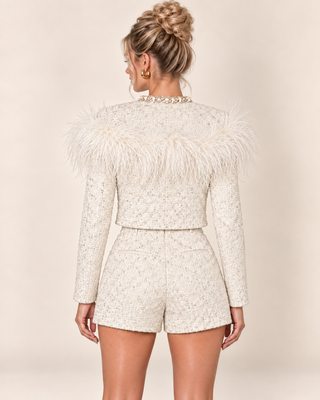A woman shown from the back wearing a cream-colored tweed jacket with dramatic feathered shoulder details and matching high-waisted shorts, set against a solid beige studio background.