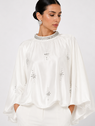 A woman wearing a white satin blouse with silver crystal embellishments on the neckline and scattered across the front, paired with white trousers against a light gray studio background.