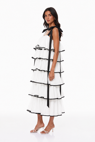 A woman modeling a white sleeveless tiered dress with black ribbon accents and black trim, paired with white heeled sandals, against a plain white studio background.