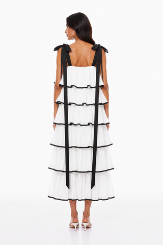 A model viewed from the back wearing a white tiered dress with black ribbon shoulder straps and black trim, paired with white heeled sandals, against a plain white studio background.