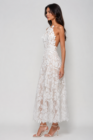 A woman modeling a white floral lace halter maxi dress with a sheer mesh overlay and an open back, paired with white strappy high-heeled sandals against a light gray studio background.