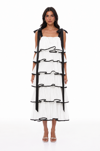 A model wearing a white tiered midi dress with black ribbon details and shoulder ties, paired with white heeled sandals, against a plain white studio background.